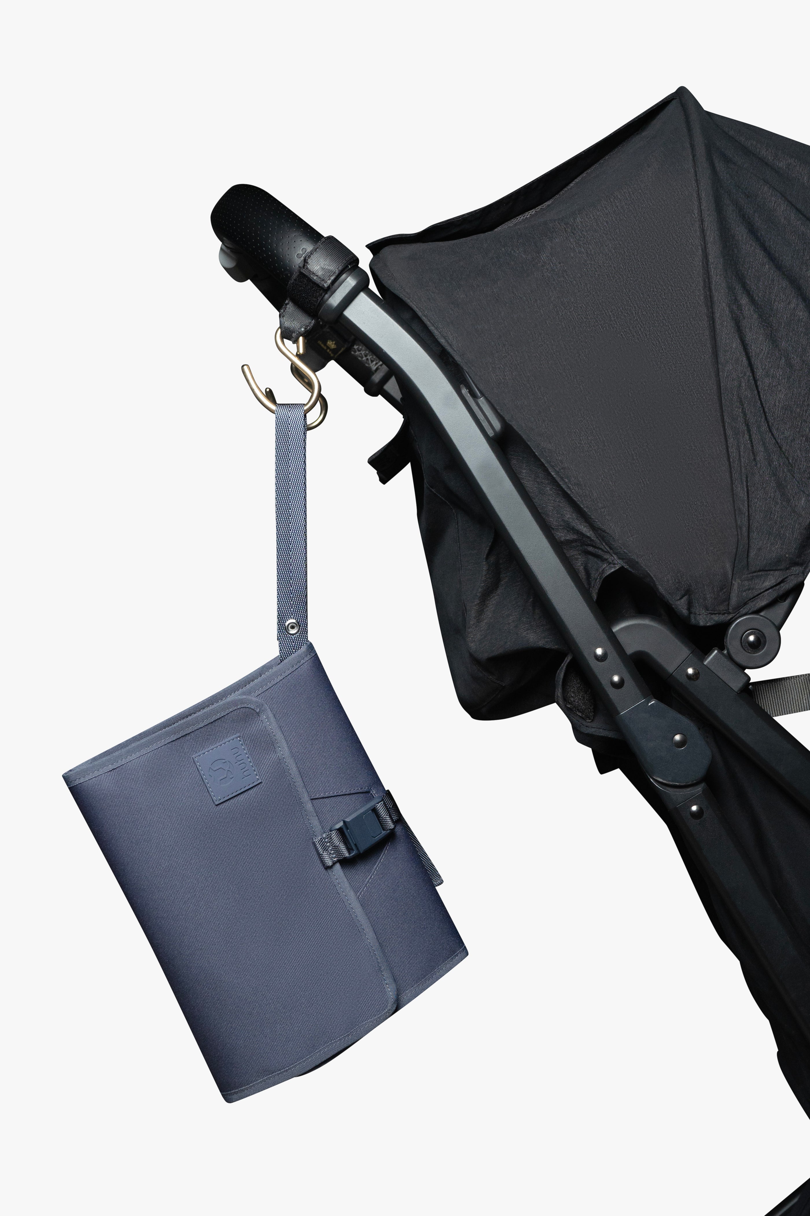 Blue changing wallet hanging from strap attached to gold stroller hook on a black stroller against white background.