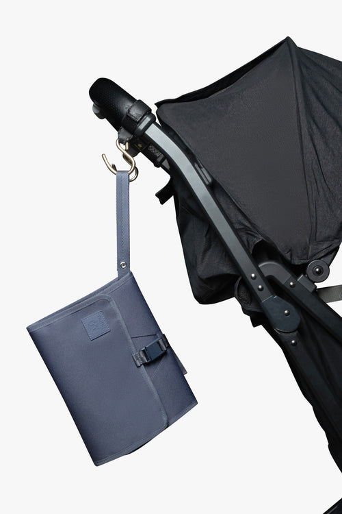 Blue changing wallet hanging from strap attached to gold stroller hook on a black stroller against white background.
