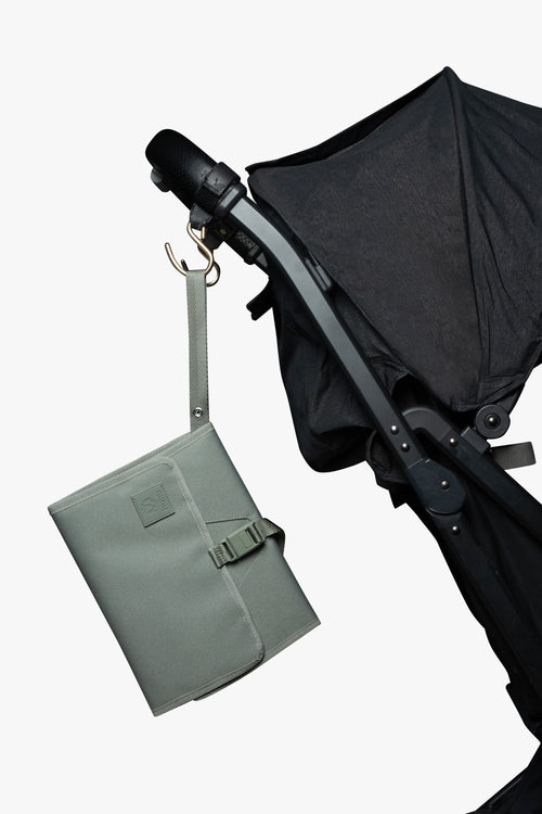 Green changing wallet hanging from strap attached to gold stroller hook on a black stroller against white background.