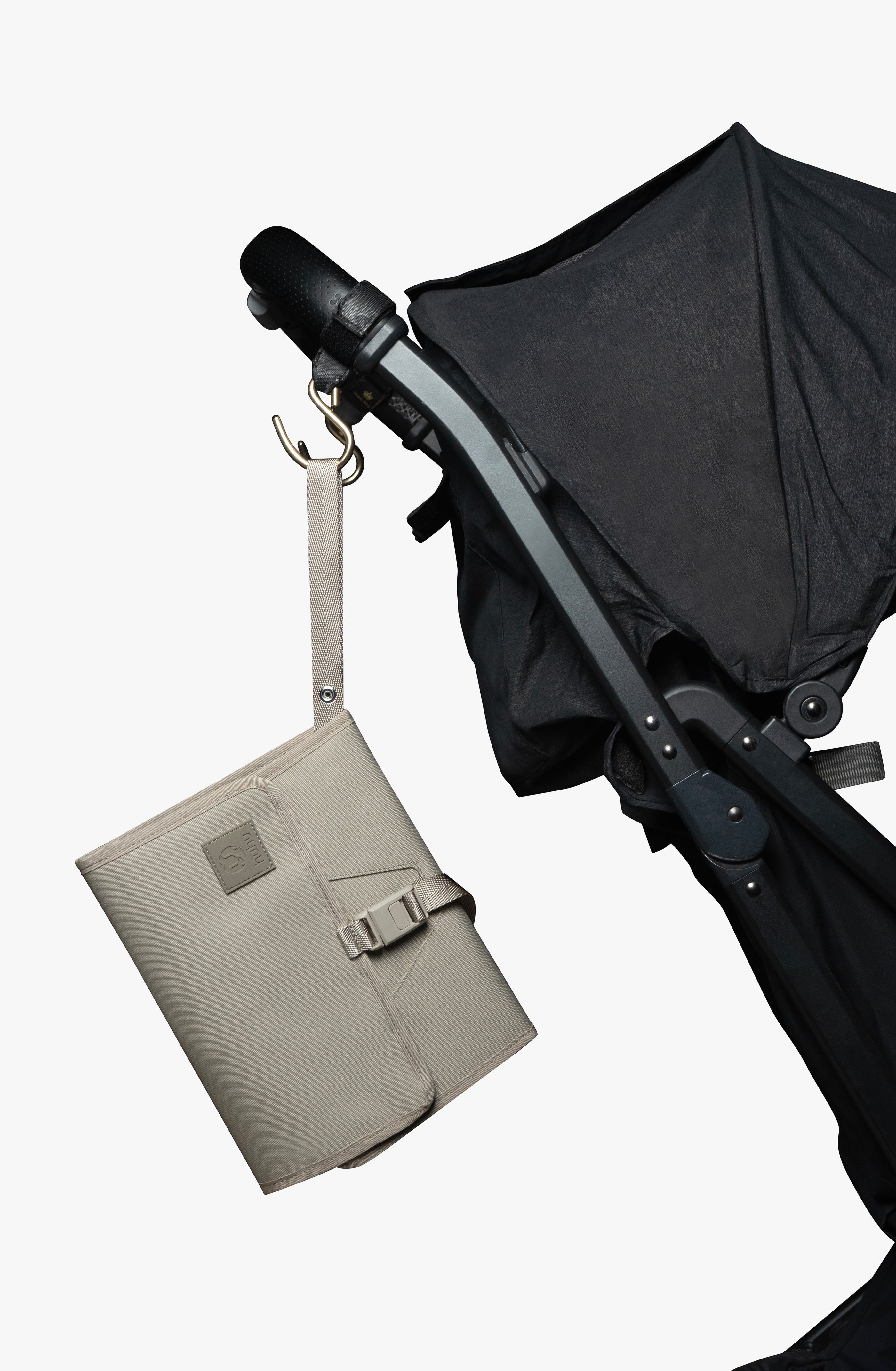 Taupe changing wallet hanging from strap attached to gold stroller hook on a black stroller against white background.