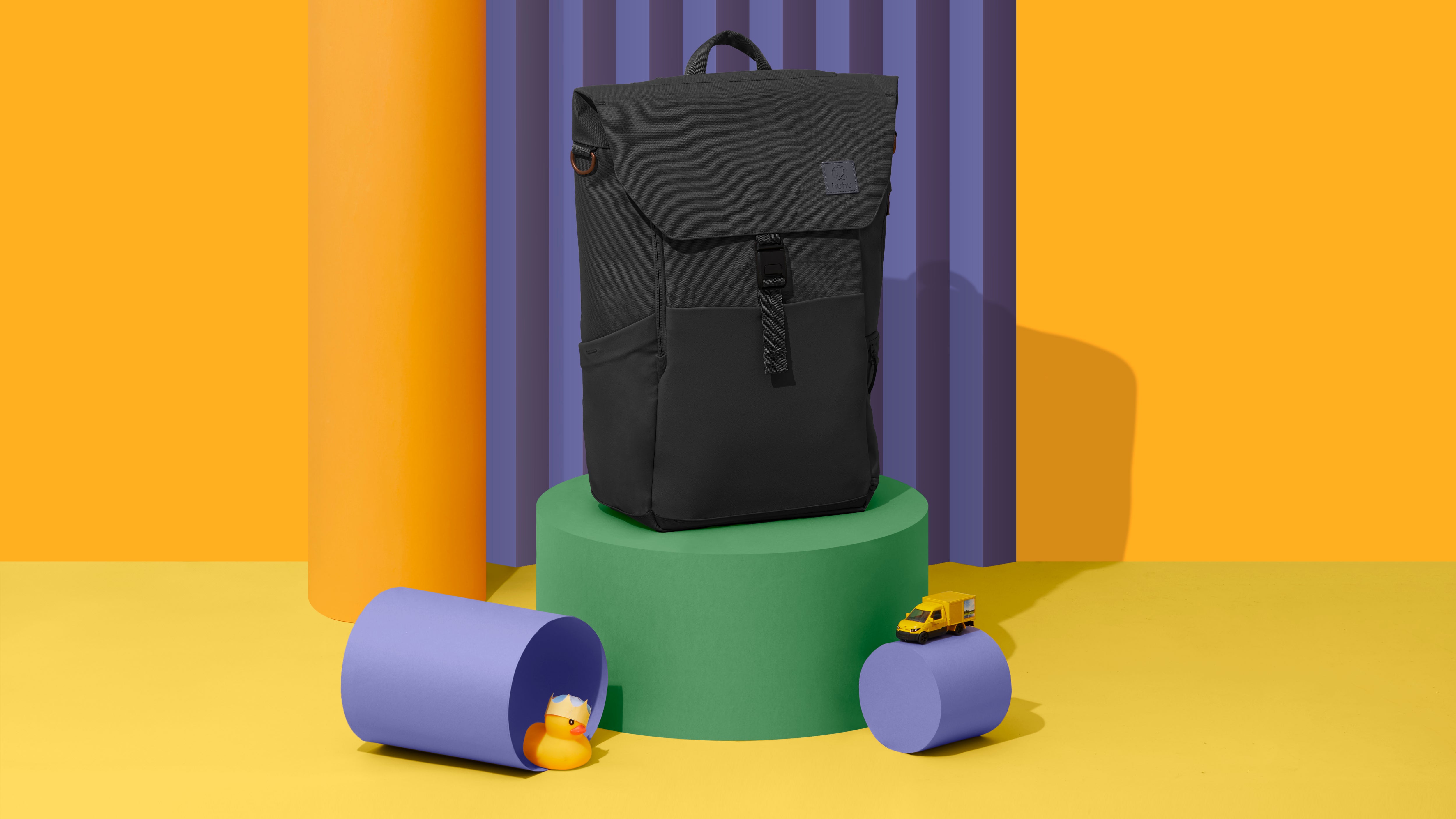picture of a black diaper backpack on a green pedestal against a yellow and purple background. There are a few props next to the backpack, specifically a yellow rubber duck and a yellow toy truck. 